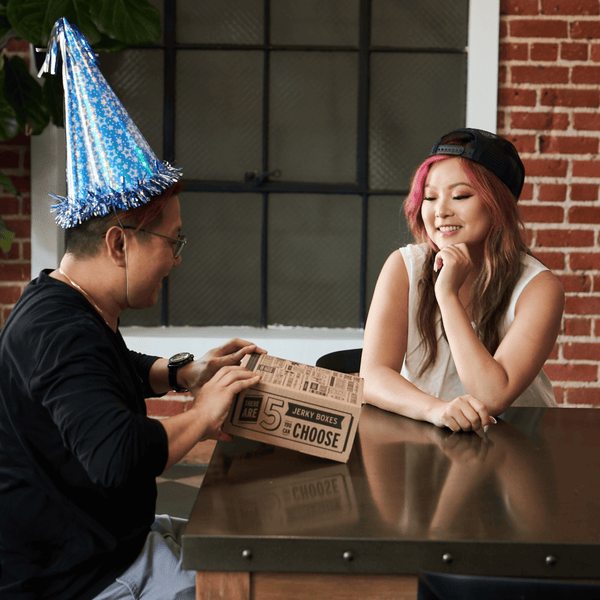 Two people seated at a table in a real indoor environment. One person gifting another person a jerky box. 