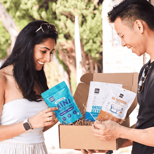 A man and a woman smiling as they open a cardboard box containing pet food bags. The woman holds a bright blue bag labeled 'Mini Beef Sticks', and the box contains other pet food bags including 'Original Beef' and 'Big Tacos'.