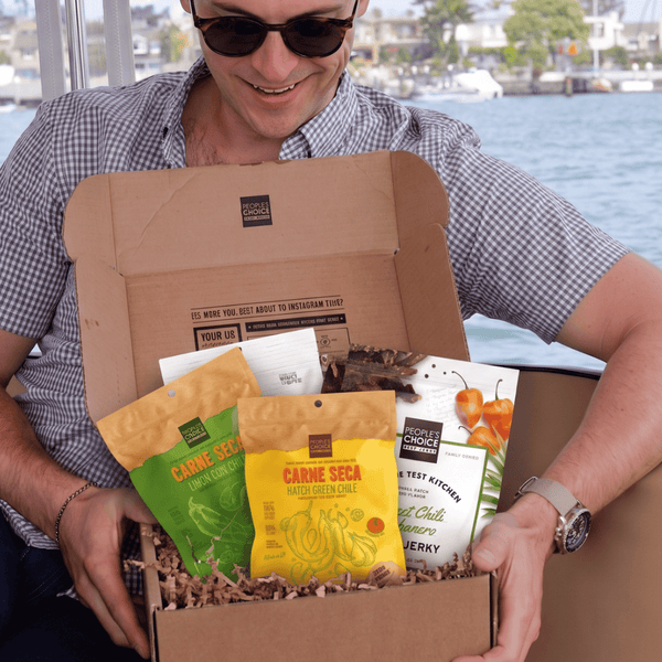 A man wearing sunglasses and a checkered shirt sits on a boat holding an open cardboard box filled with packaged snacks and coffee. Water and a distant shoreline are visible in the background.